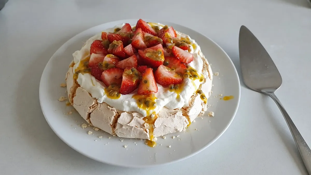 Pavlova topped with whipped cream, sliced strawberries and passionfruit pulp on a white plate