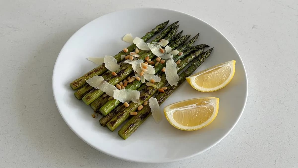 Grilled asparagus spears topped with shaved parmesan and pine nuts with lemon wedges on a white plate