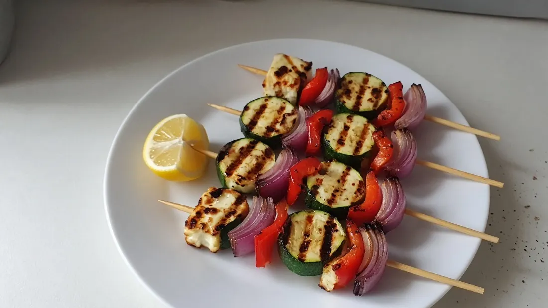 Grilled haloumi, zucchini, red capsicum and red onion skewers with a lemon wedge on a white plate