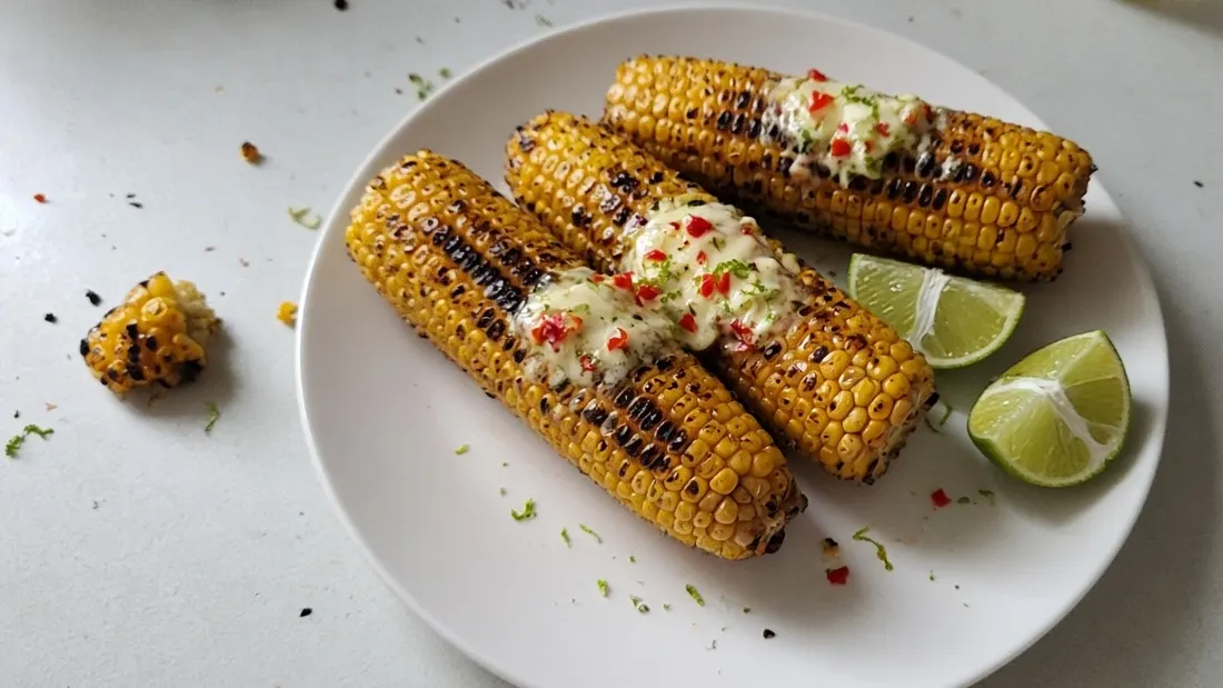Three barbecued corn cobs topped with chilli lime butter and lime wedges on a white plate