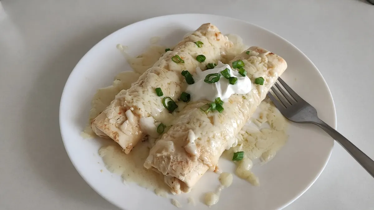 Two creamy chicken enchiladas topped with sour cream and sliced scallions on a white plate