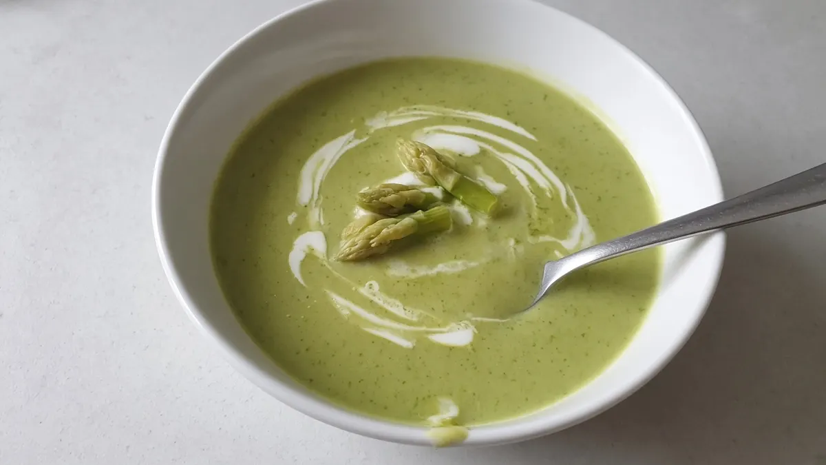 Bowl of pale green cream of asparagus soup with a swirl of cream and asparagus tips