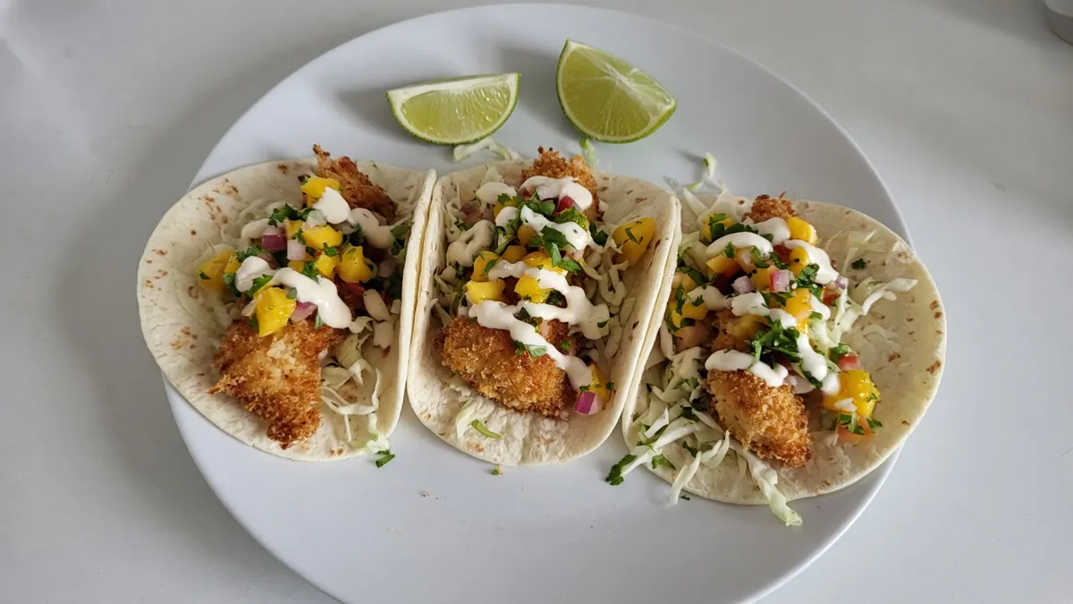 Three crumbed fish tacos with shredded cabbage, mango salsa, crema and lime wedges on a white plate