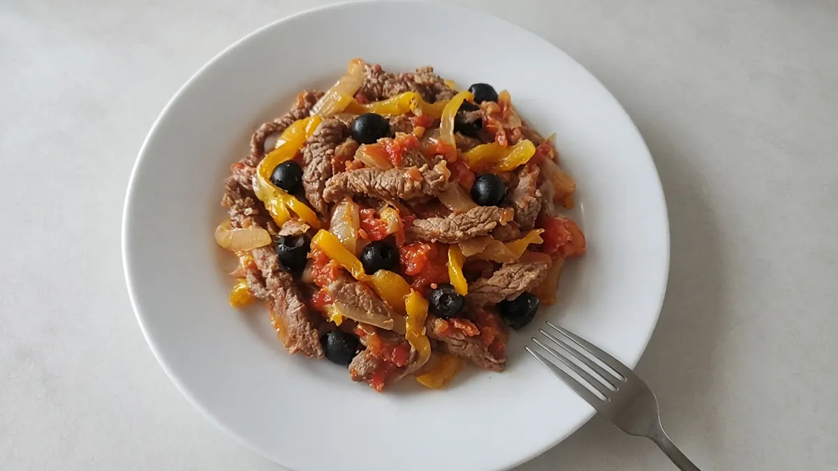 Beef strips with yellow pepper, tomatoes and black olives on a white plate with a fork