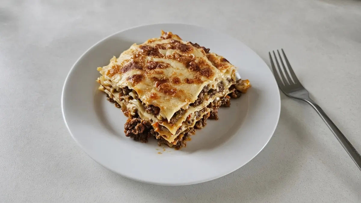 A square of beef lasagne with a golden white-sauce top on a white plate with a fork