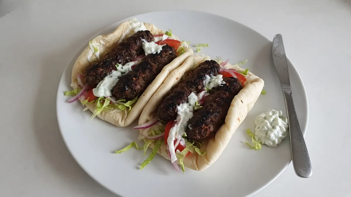 Two flatbread wraps filled with grilled lamb kofta, tzatziki, lettuce, tomato and red onion on a white plate