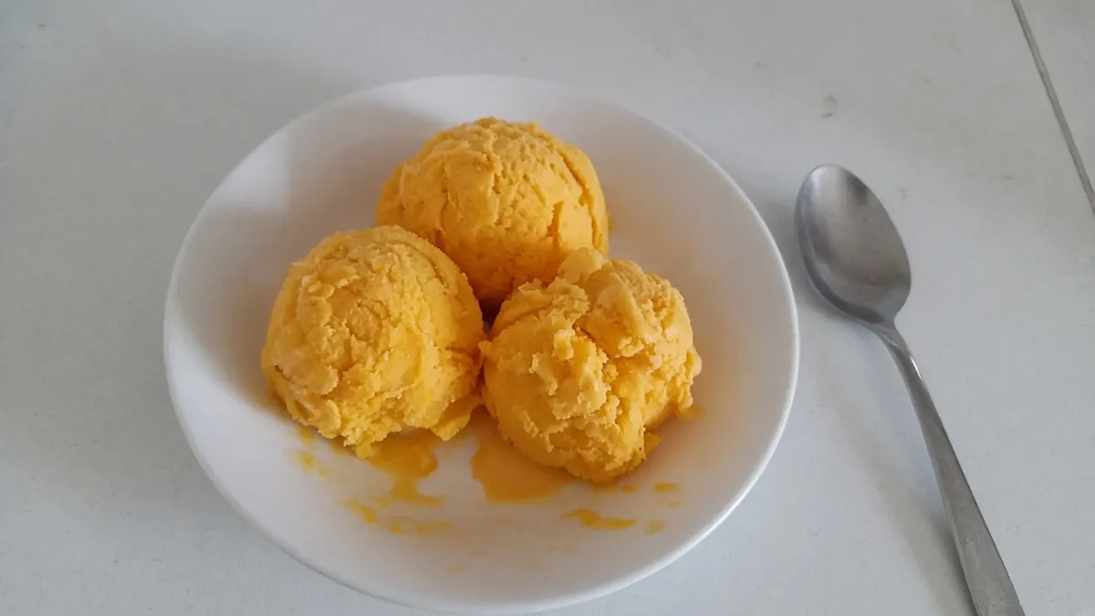 Three scoops of mango sorbet in a white bowl with a spoon beside it