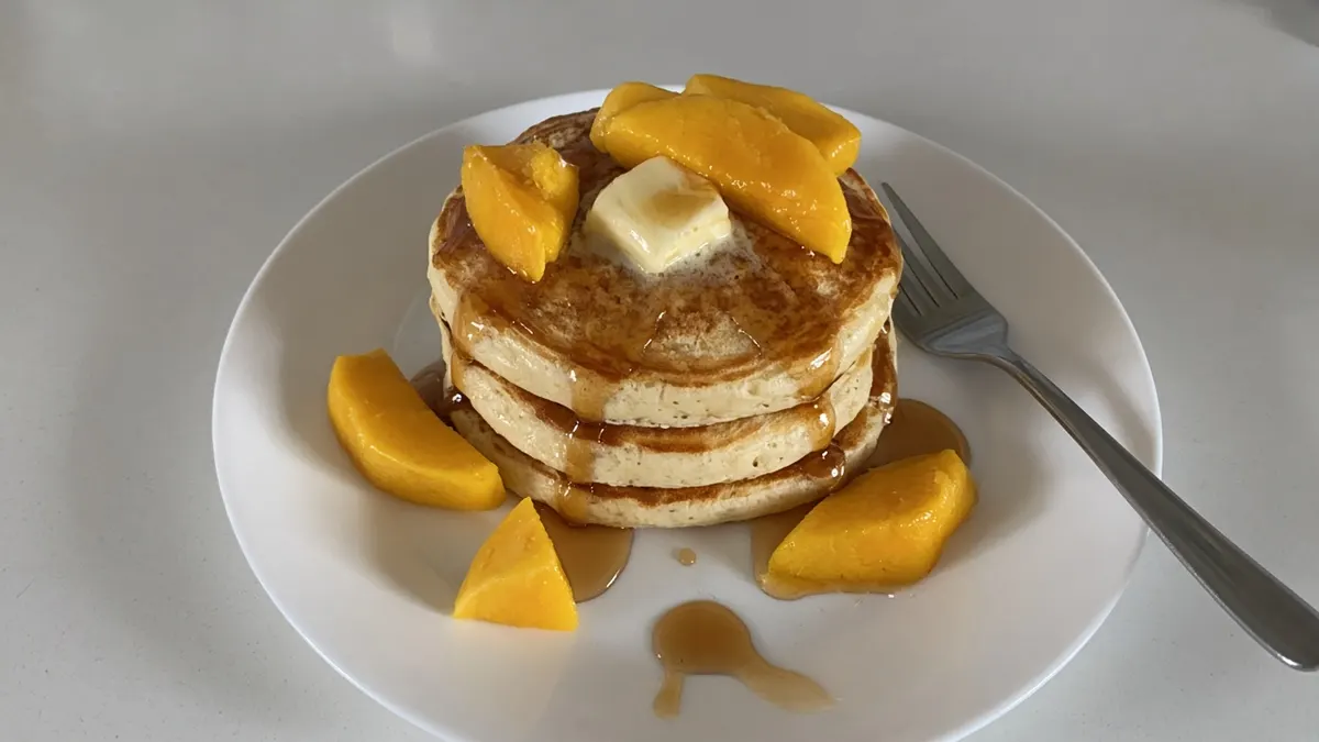 Stack of ricotta hotcakes with mango slices, a pat of butter and maple syrup on a white plate