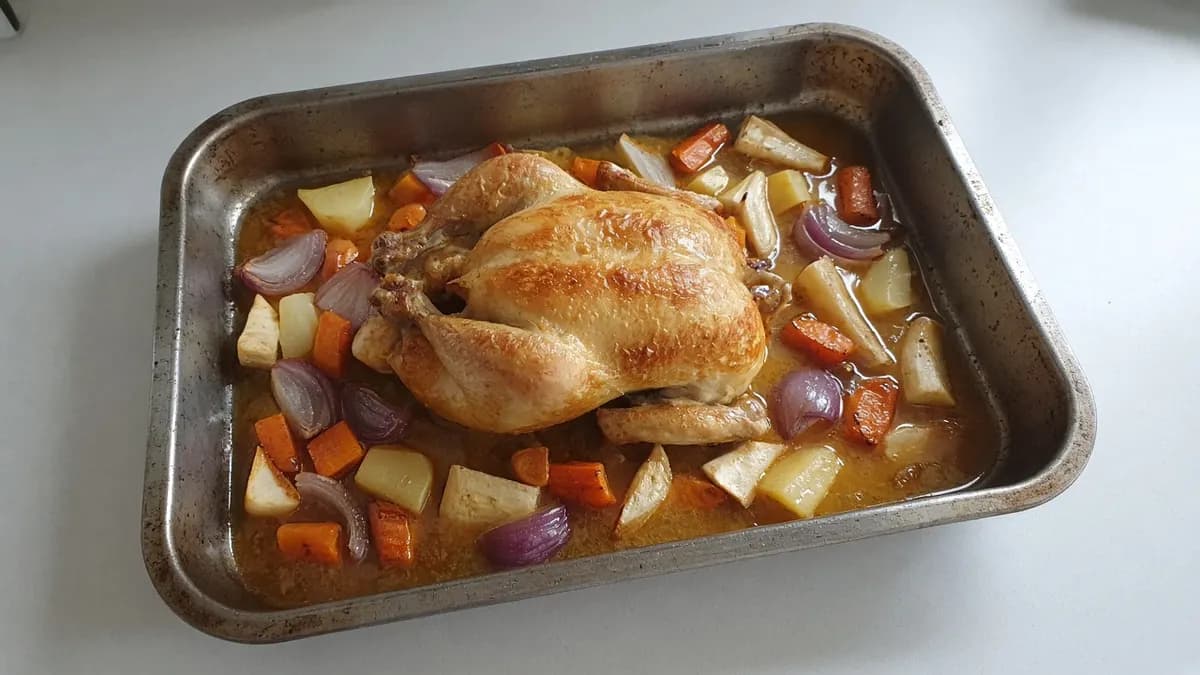 Golden whole roast chicken on carrots, potatoes and red onion in a metal roasting tray