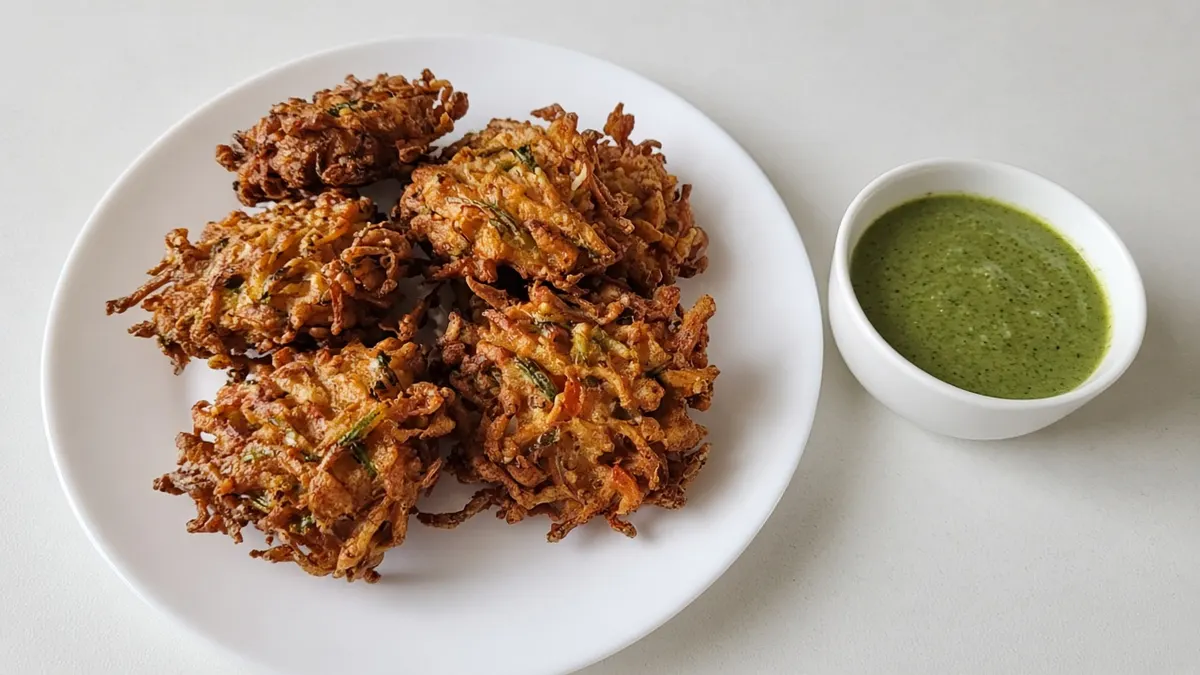Golden vegetable pakoras on a white plate with a small bowl of green coriander chutney