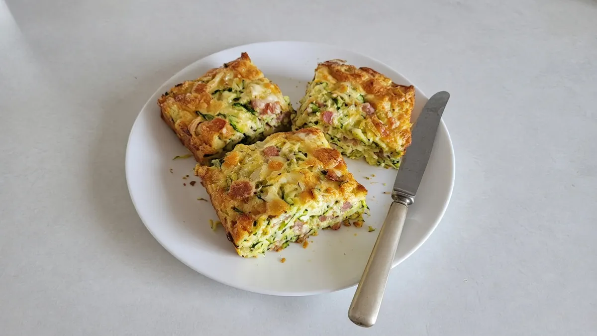 Three squares of zucchini slice with bacon and cheese on a white plate with a knife